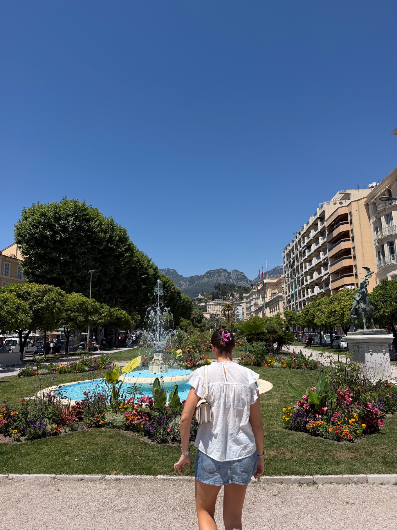 Holly walking toward a sunlit fountain and flower gardens in a grand Provençal town square, with pastel buildings and mountains rising in the distance.